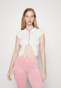 Top branco corto com um acabamento em ondas e mangas curtas, apresentando uma frente abotoada. Combinado com calças de veludo rosa.