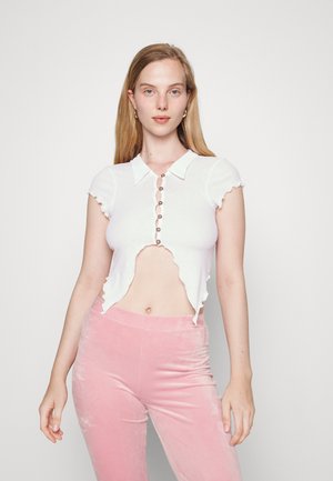 Top branco corto com um acabamento em ondas e mangas curtas, apresentando uma frente abotoada. Combinado com calças de veludo rosa.