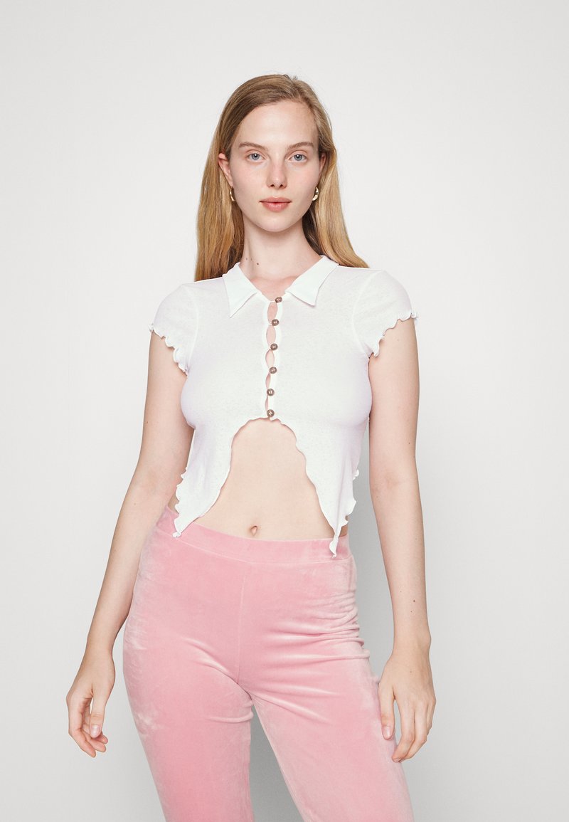 Top branco corto com um acabamento em ondas e mangas curtas, apresentando uma frente abotoada. Combinado com calças de veludo rosa.