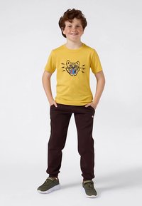 Berniukas su garbanotais raudonais plaukais stovi šypsodamasis, rankas kišenėse, vilkintis geltoną marškinėlį su riaumojančio katino grafika, tamsias kelnes ir žalias sportinius batelius.