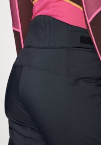 Pantaloni sportivi neri con una texture liscia, dotati di un ampio cinturino in vita e un dettaglio in Velcro. Il primo strato è abbinato a una maglietta rosa, parzialmente visibile.