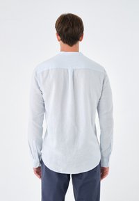 Camicia a maniche lunghe blu chiaro in lino, con colletto a bottoni e orlo curvo. La vista posteriore mostra una texture liscia e un design semplice.