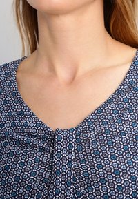 Halenka s krátkými rukávy v námořnické modři s geometrickým vzorem v bílé a aqua. Disponuje kulatým výstřihem a na středu má nařasené detaily.