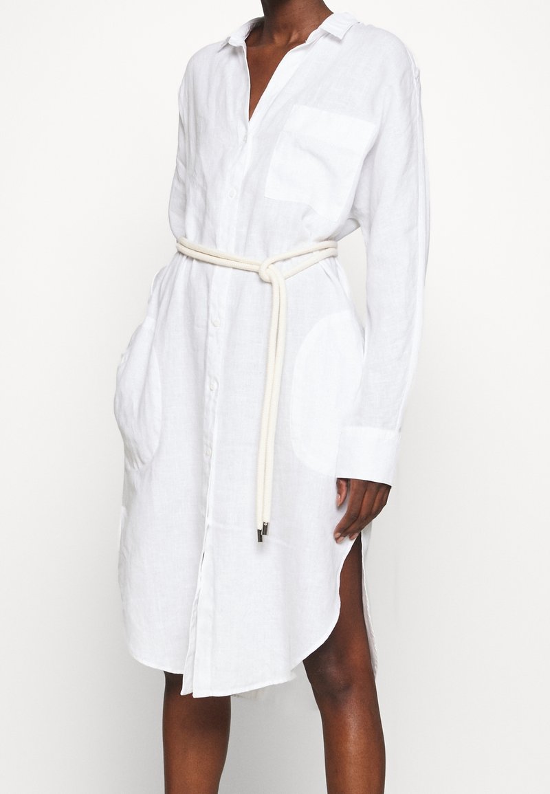 Una persona che indossa un lungo vestito bianco a camicia con cintura di corda annodata, dotato di tasche laterali e colletto leggermente aperto.
