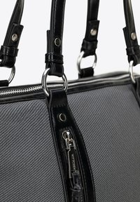 Šedá textilní kabelka s černými koženými doplňky, která má stříbrné kovové detaily, dvojité ucha a zapínání na zip. Jsou viditelné polstrované struktury.