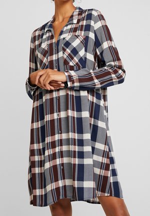 Vrouw draagt een lange mouw, blousejurk met knopen en een marineblauw, bruin en wit geruit patroon, staand met de handen geklemd ter hoogte van de taille.