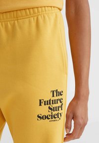 Gula sweatpants i bomull, med ett svart grafiskt tryck på vänster ben som säger "The Future Surf Society O'Neill."