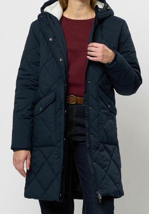 Osoba w granatowym pikowanym długim płaszczu z kapturem, bordowym swetrze, ciemnych dżinsach i brązowym pasku, trzymająca miejsce zamka płaszcza.