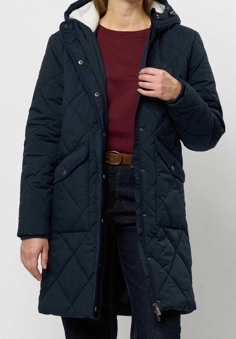 Persona que lleva un abrigo largo acolchado azul marino con capucha, suéter granate, vaqueros oscuros y cinturón marrón, sujetando la zona de la cremallera del abrigo.