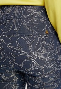 Tummansiniset housut, joissa on valkoinen kukkakuosi, takatasku ja pieni kultalogo. Kangas näyttää teksturoidulta ja denimin kaltaiselta kudokselta.