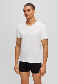 T-shirt blanc à manches courtes en coton lisse, avec un col rond et un petit logo de marque sur la poitrine gauche.
