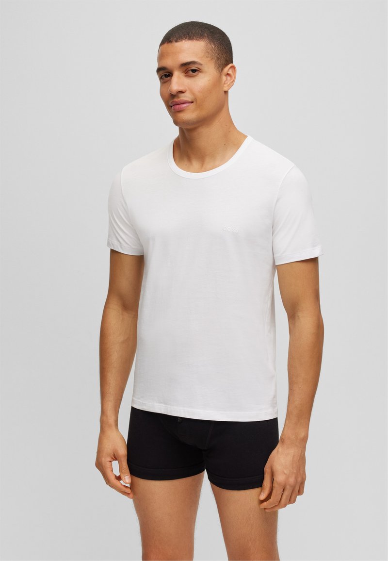 T-shirt blanc à manches courtes en coton lisse, avec un col rond et un petit logo de marque sur la poitrine gauche.
