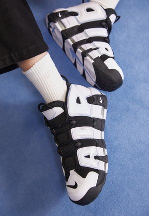 Stopy w czarno-białych sneakersach Nike Air More Uptempo i białych skarpetkach z prążkami na niebieskim tle.