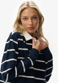 Marineblauwe trui met witte horizontale strepen, met daaronder een wit overhemd met kraag. Zachte textuur en een ontwerp met een ontspannen pasvorm.