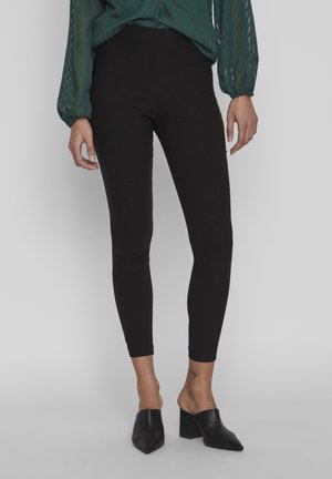 Kobieta ubrana w czarne dopasowane spodnie do kostek, ciemnozieloną bluzkę z długim rękawem oraz czarne mules na szerokim obcasie, stojąca na jednolitym tle.