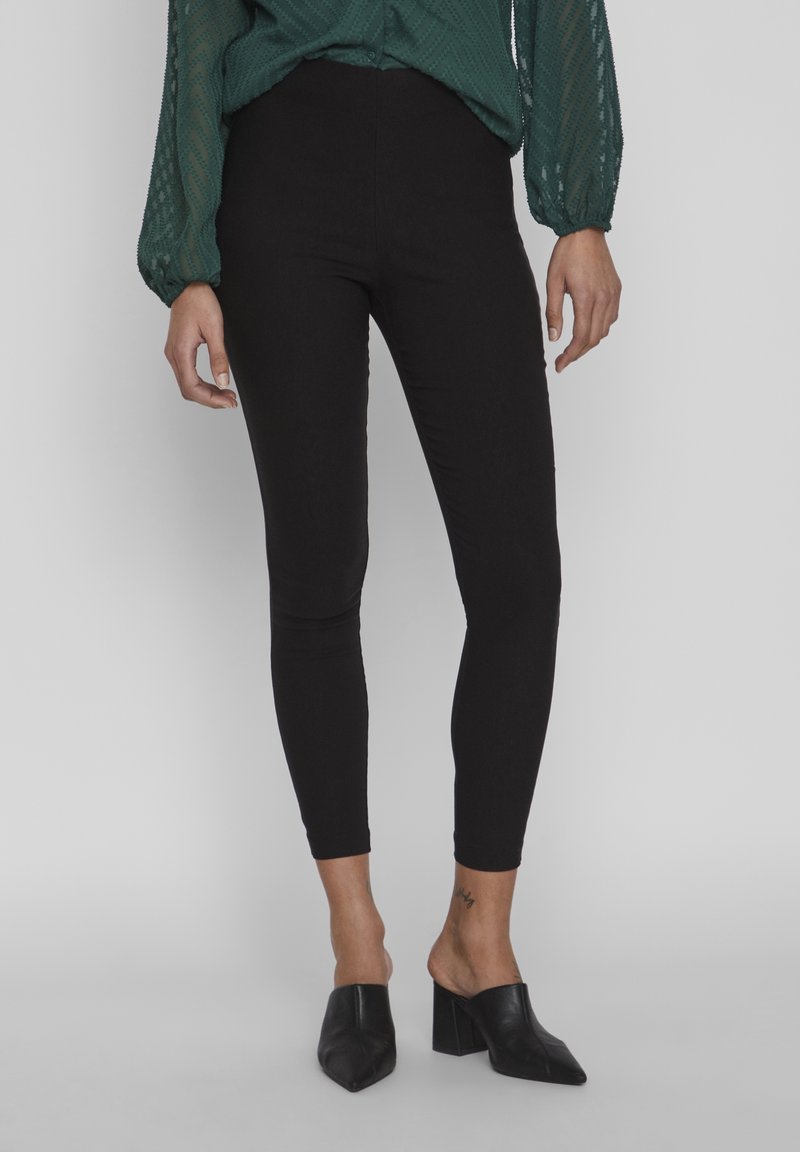 Femme portant un pantalon noir ajusté aux chevilles, un chemisier vert foncé à manches longues et des mules noires à talons blocs, debout devant un fond uni.