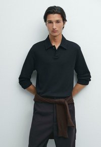 Svart långärmad pikétröja med strukturerad finish, med krage och placket med tre knappar, ihopparad med mörka byxor och en brun tröja knuten runt midjan.