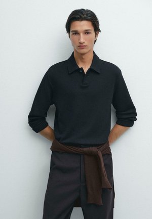 Zwarte longsleeve polo met een gestructureerde afwerking, voorzien van een kraag en een knoopsluiting met drie knopen, gecombineerd met donkere broek en een bruine trui om de taille geknoopt.