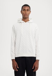 Bluza cu glugă de culoare deschisă, cu un model repetitiv de litere stilizate. Material din bumbac, croială lejeră și detalii cu șnur.