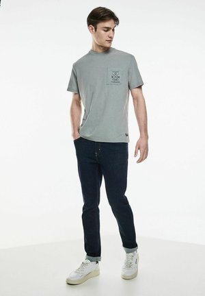 Gray cotton t-shirt with a round neck and short sleeves, featuring a chest pocket with black graphic print. Worn with dark jeans and white sneakers.