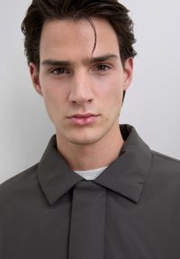 Jonge man met donker haar, gekleed in een donkergrijze colbert over een wit shirt, recht vooruit kijkend met een neutrale uitdrukking voor een effen achtergrond.