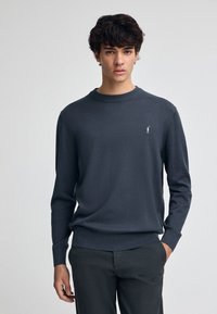 Pull noir à manches longues avec un col rond, des poignets et un ourlet côtelés, arborant un petit logo blanc sur la zone gauche de la poitrine.