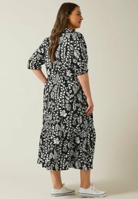 Schwarzes Blumen-Midikleid mit weißen botanischen Druckmotiven, kurzen aufgerollten Ärmeln, einer taillierten Taille und gewelltem Saum. Kombiniert mit weißen Sneakers.