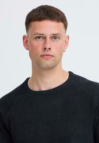 Zwarte geribde gebreide trui met een ronde hals en raglan mouwen. Model toont een neutrale uitdrukking. Een gouden ringoorbel is zichtbaar.