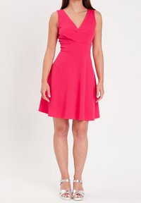 Vestido rosa sin mangas con escote en V y cintura ajustada, acampanado en el dobladillo. Modelo con sandalias de tacón plateadas y tiras.