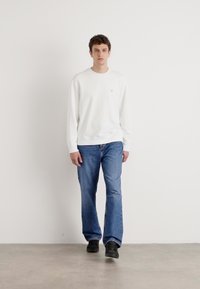 Sudadera blanca de manga larga, vaqueros azul, zapatos negros. Ajuste casual con puños y dobladillo acanalados. El modelo se encuentra de pie frente a un fondo simple.