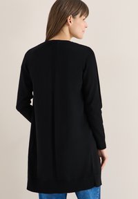 Femme de profil portant un long cardigan noir à bordures côtelées sur un jean bleu, sur un fond beige uni.
