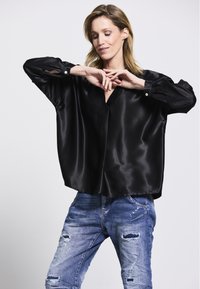 Schwarze Satin-Bluse mit V-Ausschnitt und lockerer Passform, ausgestattet mit langen Ärmeln mit Knopf-Akzenten; kombiniert mit zerrissenen Jeans.
