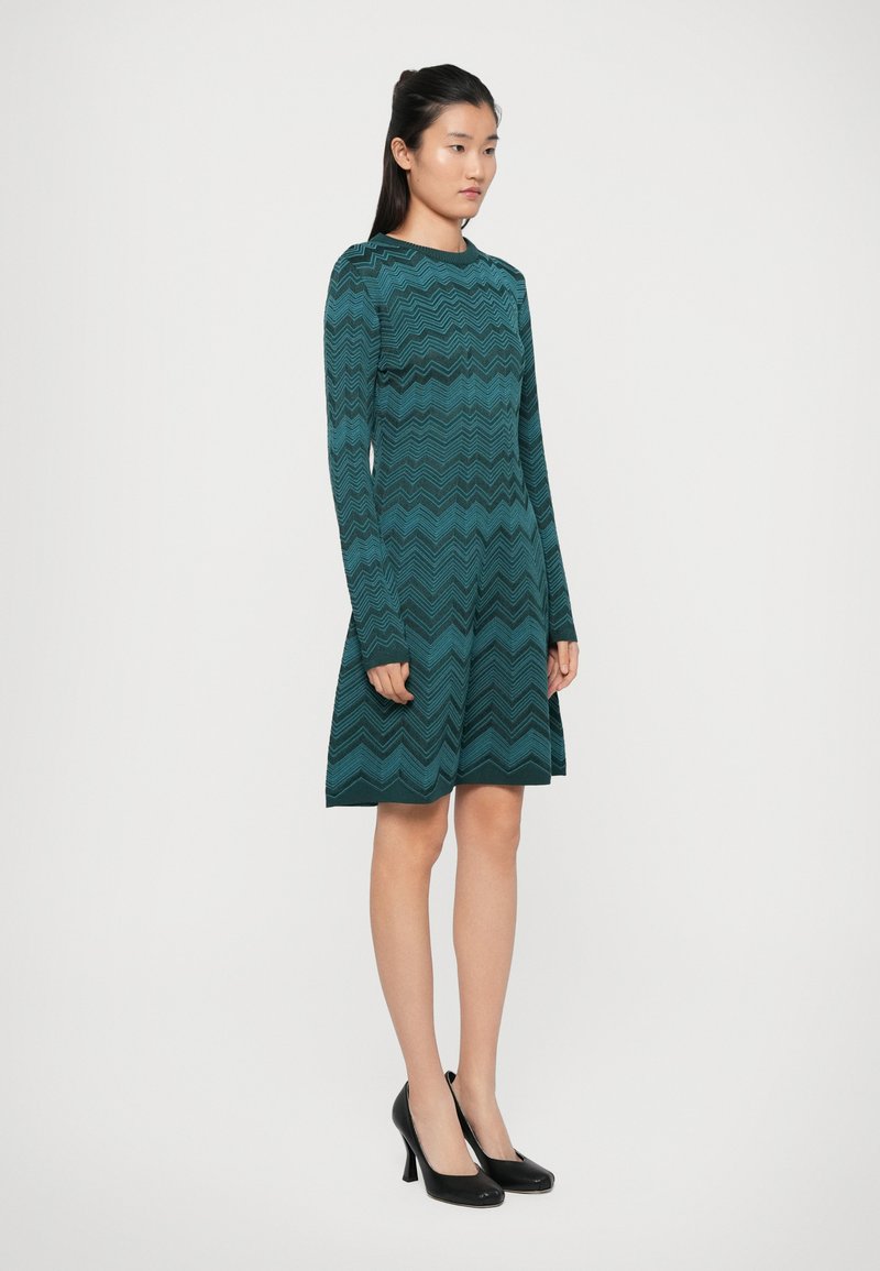 Robe longue teal à manches longues avec un motif en zigzag, corsage ajusté, jupe évasée et col rond, associée à des chaussures à talons hauts noires.