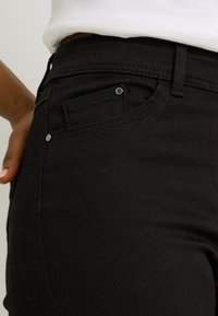 Jeans de mezclilla negra con un corte ajustado, que presentan un bolsillo frontal, costuras en contraste y un detalle de botón metálico en la cintura.