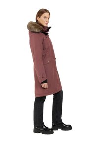 Parka lunga borgogna con cappuccio rifinito in pelliccia sintetica, chiusura con zip e bottoni a pressione, due tasche frontali, indossata su jeans neri e stivali neri massicci.