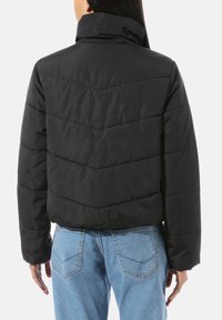 Czarna kurtka puchowa z przeszyciami w kształcie litery V, wysokim kołnierzem i luźnym krojem. Noszona z jasnoniebieskimi dżinsami. Długie rękawy i gładka faktura.