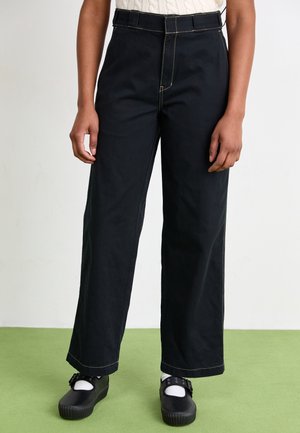 Person, die schwarze Hosen mit weitem Bein und sichtbaren beigen Nähten, weiße Socken und schwarze Riemenschuhe trägt, steht auf grünem Boden.