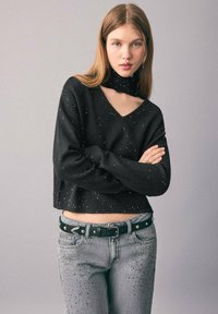 Svart kortärmad tröja med V-hals och matchande hög hals, dekorerad med små prickar. Bärs med grå slitna jeans och ett svart bälte.