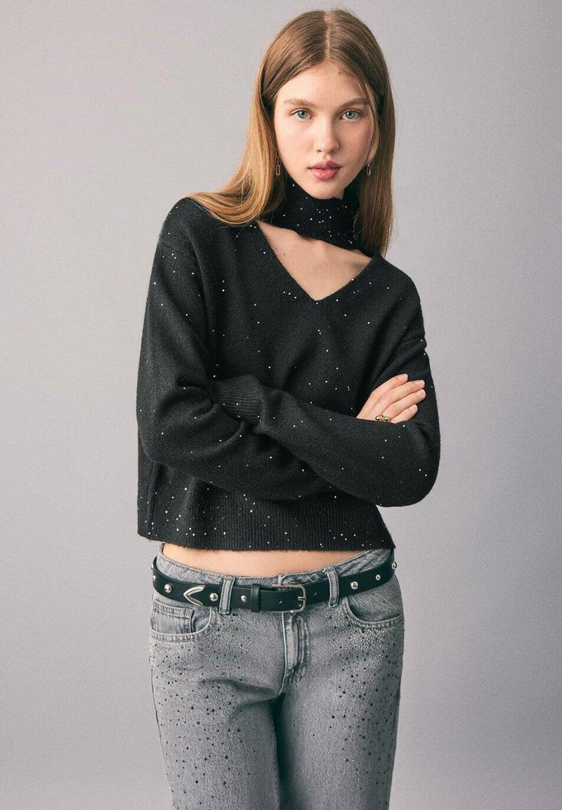 Svart kortärmad tröja med V-hals och matchande hög hals, dekorerad med små prickar. Bärs med grå slitna jeans och ett svart bälte.