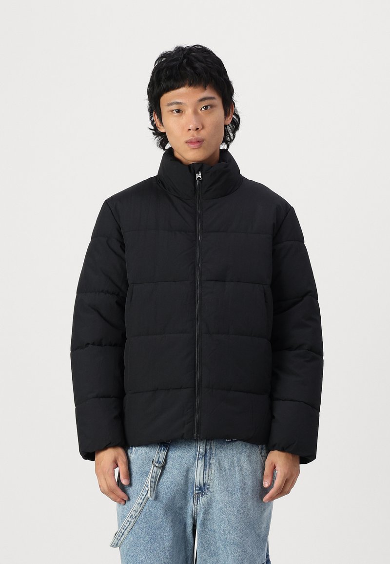 Black puffer jacket with a high collar, featuring quilted sections, front zipper, and a relaxed fit. Worn over light blue jeans.