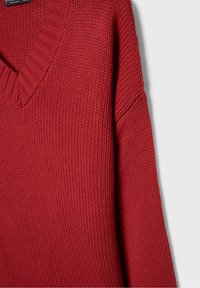 Rode gebreide trui met een V-hals ontwerp, voorzien van een gestructureerd patroon en verlaagde schouders. De stof lijkt zacht en dik.