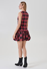 Donna che indossa un mini abito senza maniche a quadretti rosso e nero con gonna a ruota e stivali neri alla caviglia, in piedi di profilo su sfondo bianco.