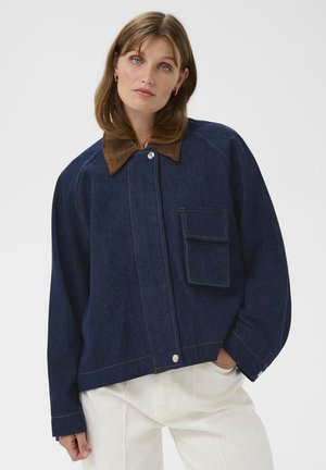 Vrouw draagt een donkerblauwe spijkerjas met een bruine kraag, een oversized zak en witte broek, staand tegen een effen achtergrond.