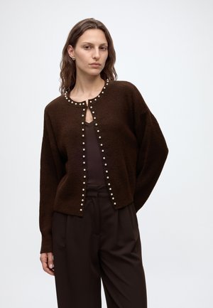 Femme portant un cardigan marron foncé avec des boutons en perle sur un haut foncé et un pantalon assorti, debout devant un fond clair uni.