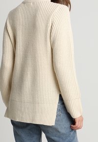 Person, der einen cremefarbenen Grobstrickpullover mit seitlichen Schlitzen und lässigen blauen Jeans vor einem schlichten, hellen Hintergrund trägt.