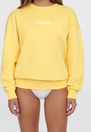 Nainen, jolla on päällään löysä keltainen collegepaita, jossa on teksti "The Future Surf Society" rinnassa, ja valkoiset bikinihousut. Hän seisoo yksiväristä taustaa vasten.