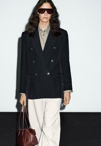Person mit langen dunklen Haaren, die ein übergroßes schwarzes zweireihiges Blazer, ein beiges Hemd, weiße Hosen, dunkle Sonnenbrille trägt und eine braune Ledertasche hält.