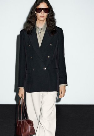 Person with long dark hair wearing oversized black double-breasted blazer, beige shirt, white pants, dark sunglasses, carrying brown leather handbag.