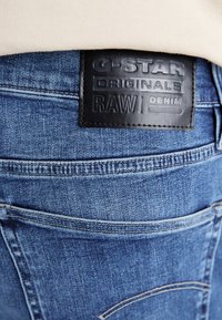Blå denimjeans med en texturerad yta, som har en läderlapp med texten "G-Star Originals RAW Denim" och synliga sömnadsdetaljer.