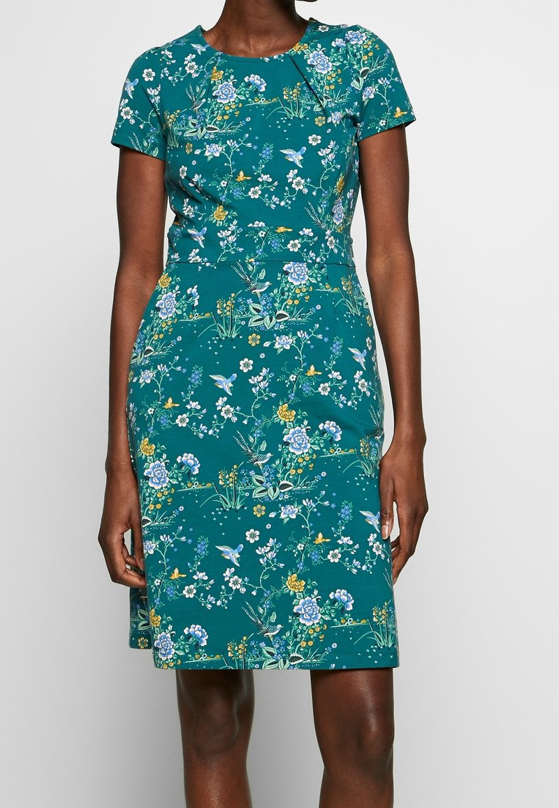 Robe courte à manches courtes bleu sarcelle avec imprimé floral et oiseaux, taille cintrée, portée par une personne debout sur un fond uni.